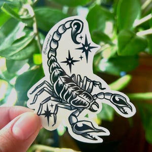May include: A die-cut sticker featuring a black and white illustration of a scorpion with a curved tail and pincers. The design includes three four-pointed star shapes with small hearts in the centre and the text @illustratedmd on the side.