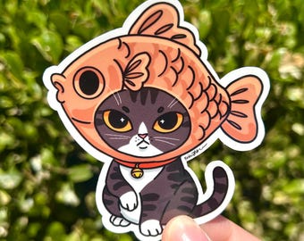 May include: A die-cut sticker featuring a dark gray and white tabby cat wearing a peach-colored fish-shaped hood. The cat has large amber eyes and a small gold bell on a red collar. The illustration has a white border and a signature on the side.