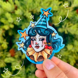 May include: A sticker depicting a stylized clown with teal hair, a party hat, and star accents. The clown has red cherry earrings and a ruffled collar. The sticker is held by a hand against a green background.