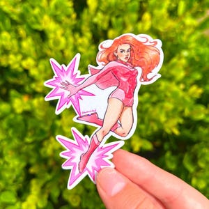 May include: A sticker featuring a superheroine with red hair and a pink costume, cape, and boots. She is depicted in a dynamic pose with pink energy bursts emanating from her hands and feet. The sticker has a white border.