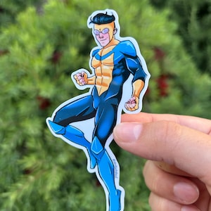 May include: A sticker featuring a comic book style illustration of a superhero in a blue and black suit with yellow accents. The character is in a dynamic pose, with a determined expression. The sticker is held against a backdrop of green foliage.