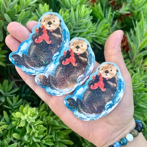 Op de afbeelding: Drie stickers met een zeeotter die een rode zeester vasthoudt. De stickers hebben een blauwe rand en een glittereffect. De zeeotters zijn bruin en zwart, met bruine gezichten. De stickers worden in een hand gehouden.