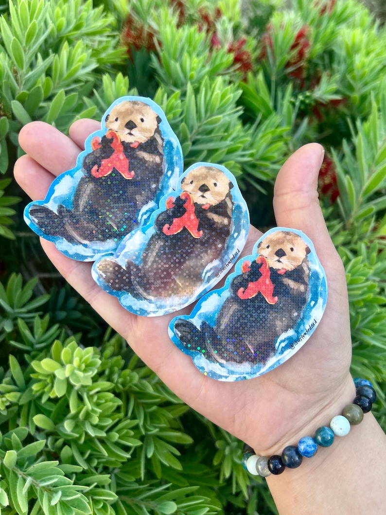 Op de afbeelding: Drie stickers met een zeeotter die een rode zeester vasthoudt. De stickers hebben een blauwe rand en een glittereffect. De zeeotters zijn bruin en zwart, met bruine gezichten. De stickers worden in een hand gehouden.