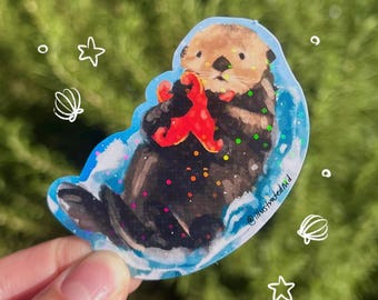 Puede incluir: Una pegatina troquelada que muestra una nutria marina marrón flotando en agua azul mientras sostiene una estrella de mar roja brillante. La ilustración incluye acentos de purpurina coloridos y garabatos blancos de estrellas y conchas, con el texto @illustratedmd.