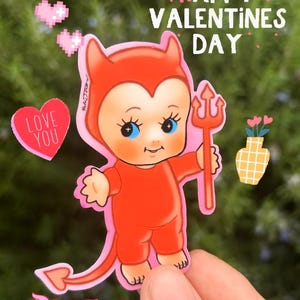 Könnte beinhalten: Ein Aufkleber mit einem Cartoon-Teufel in roter Kleidung, der einen Dreizack hält. Der Aufkleber hat den Text "HAPPY VALENTINES DAY" und "LOVE YOU" mit rosa Herzen und einer kleinen Blumenvase. Die Figur hat blaue Augen.