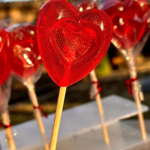 May include: A close-up of red heart-shaped lollipops on sticks. The lollipops are arranged on a white tiered stand.