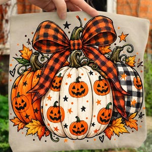 May include: A beige tote bag featuring a graphic print of three pumpkins with jack-o'-lantern faces. The pumpkins are decorated with black and orange stars and tied together with a large orange and black buffalo plaid ribbon bow.