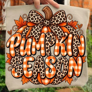 May include: A beige t-shirt featuring a large graphic of a pumpkin with a leopard print bow. The pumpkin has orange and white checkered patterns. The text PUMPKIN SEASON is written in bold, orange and white checkered letters with leopard print accents.