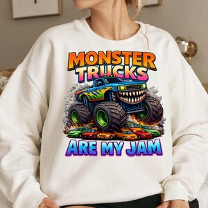 Puede incluir: Sudadera blanca con un gr&aacute;fico colorido de un monster truck con una sonrisa, llamas y el texto "MONSTER TRUCKS ARE MY JAM". El monster truck es azul con detalles verdes y amarillos, y las ruedas son verdes.