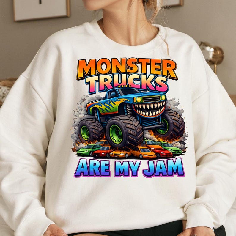 Puede incluir: Sudadera blanca con un gr&aacute;fico colorido de un monster truck con una sonrisa, llamas y el texto "MONSTER TRUCKS ARE MY JAM". El monster truck es azul con detalles verdes y amarillos, y las ruedas son verdes.