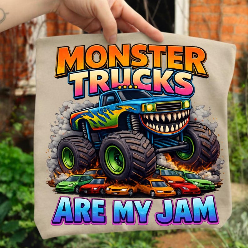 Puede incluir: Un bolso tote beige con un gr&aacute;fico colorido de un cami&oacute;n monstruo azul con una sonrisa dentada, llamas y neum&aacute;ticos grandes. El texto "MONSTER TRUCKS ARE MY JAM" se muestra en colores vibrantes arriba y abajo del cami&oacute;n.