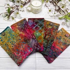 May include: Five colorful fabric cases with unique batik patterns in shades of blue, orange, red, and green. Each case has a decorative button and a brown trim. The cases are arranged on a white wooden surface.