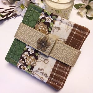 May include: A square, quilted coaster with a floral and plaid pattern in shades of green, brown, and cream. A beige strap with a flower-shaped button secures the coaster. The coaster is approximately 4 inches square.
