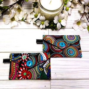 May include: Two small, colorful fabric wallets with black accents and button closures. The wallets feature vibrant patterns of circles and floral designs in shades of red, orange, yellow, blue, and green against a black background. A black strap is attached to each wallet.