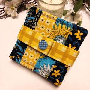 May include: A square, quilted fabric item with a yellow gingham strap and a blue button closure. The patchwork design features yellow daisies, sunflowers, and blue and white floral patterns on a black background. The item is likely a coaster or small mat.