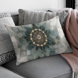 May include: A gray sofa with decorative pillows and a pink throw blanket. The rectangular pillow features a teal and gold floral design. The throw blanket is pink with a herringbone pattern and white fringe.