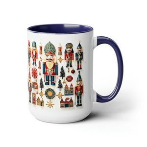 May include: White ceramic coffee mug with a blue rim and handle. The mug features a colorful pattern of nutcracker figurines, Christmas trees, stars, and other holiday decorations.