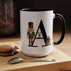 May include: White ceramic mug with a black rim and handle. The mug features a black letter "A" with a nutcracker, a reindeer, and a gift box inside the letter.