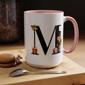 May include: White ceramic mug with a pink rim and handle. The mug features a black letter "M" with a Christmas-themed design including a nutcracker, a gift, and bells.