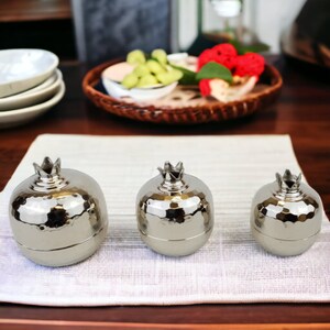 May include: Three silver metal pomegranate shaped containers with a hammered finish. The containers are of different sizes and are arranged in a row on a white textured surface.