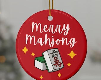 Puede incluir: Adorno navideño redondo rojo con las palabras "Merry Mahjong" en escritura blanca. El adorno presenta un gráfico de dos fichas de mahjong y estrellas amarillas. Una cuerda dorada está unida para colgar.
