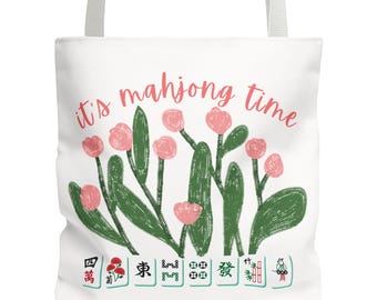 May include: White tote bag with a floral design and the text "it's mahjong time" in pink. The design includes pink flowers, green leaves, and Mahjong tiles at the bottom. The bag has white handles.