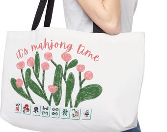 以下が含まれることがあります： 黒いストラップが付いた白いトートバッグ。「it's mahjong time」というピンク色の文字、ピンクの花と緑の葉のフローラルデザイン、下部に麻雀牌が描かれています。