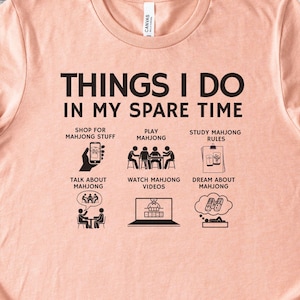 May include: A peach-colored t-shirt with the text "THINGS I DO IN MY SPARE TIME" and illustrations of mahjong-related activities. The shirt is paired with blue jeans and accessories.
