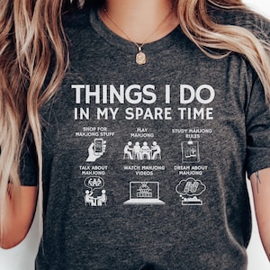 May include: Dark gray t-shirt with white text and illustrations. The shirt reads "THINGS I DO IN MY SPARE TIME" with images depicting activities like playing mahjong, studying rules, and watching videos. A fun, casual tee.