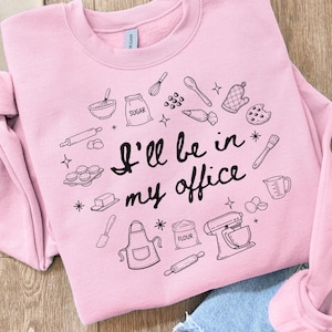 May include: A light pink sweatshirt with the phrase "I'll be in my office" in black script. The sweatshirt is decorated with black line drawings of baking tools and ingredients, including a mixer, rolling pin, and measuring cup.