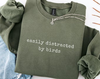 May include: A military green crewneck sweatshirt featuring the white embroidered text "easily distracted by birds" on the chest. The garment has long sleeves with ribbed cuffs and a silver-toned chain necklace resting on the neckline.