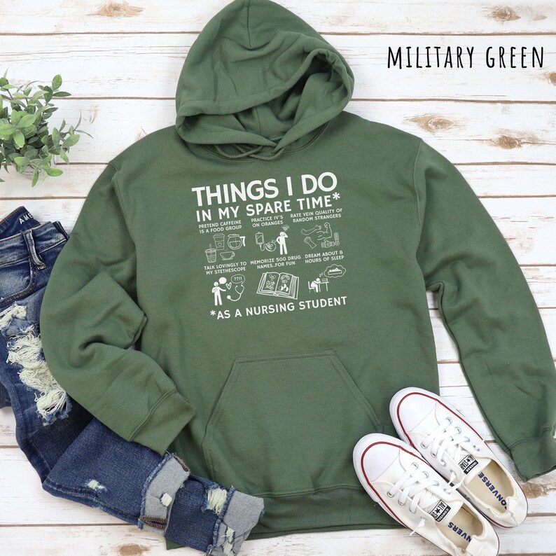 Peut inclure: Un sweat &agrave; capuche vert militaire avec le texte "THINGS I DO IN MY SPARE TIME *AS A NURSING STUDENT" en blanc. Le sweat &agrave; capuche a une poche avant et une capuche. Des baskets Converse blanches et un jean bleu sont &eacute;galement dans l'image.