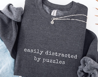 May include: Dark heather gray sweatshirt with the text "easily distracted by puzzles" in white. A silver necklace with a sunburst pendant is draped across the front. The sweatshirt is folded with a pair of blue jeans.