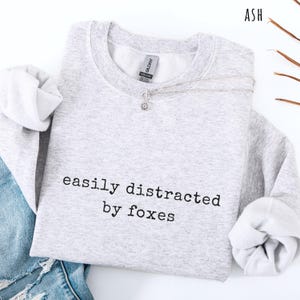 Pu&ograve; includere: Felpa grigio chiaro con la scritta nera "easily distracted by foxes". Una collana d'argento con un piccolo ciondolo &egrave; drappeggiata sulla felpa. La parola "ASH" &egrave; visibile in alto a destra.