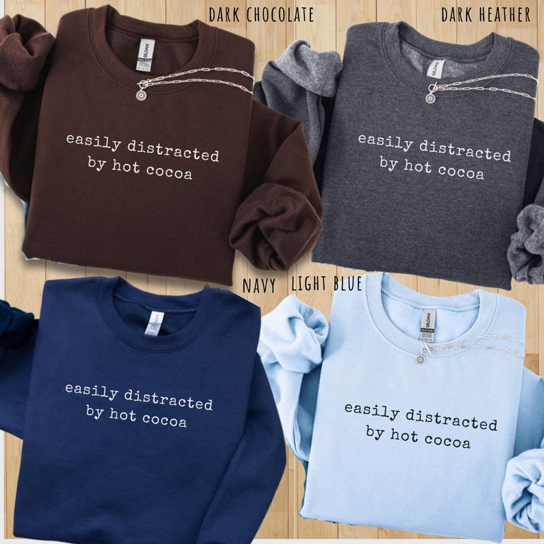 May include: Four crewneck sweatshirts in dark chocolate, dark heather, navy, and light blue. Each sweatshirt features the white text "easily distracted by hot cocoa" printed on the chest and includes a silver-toned chain necklace around the collar.