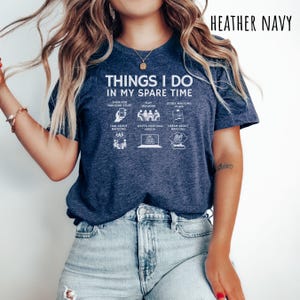 May include: Heather navy blue t-shirt with the text "THINGS I DO IN MY SPARE TIME". The graphic includes icons representing activities like shopping, playing, and studying Mahjong. The shirt is a casual wear item.
