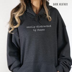 Pu&ograve; includere: Felpa con cappuccio grigio scuro con la scritta bianca "easily distracted by foxes". La felpa ha un cappuccio con coulisse e una tasca frontale. Le parole "DARK HEATHER" sono stampate nella parte superiore dell'immagine.