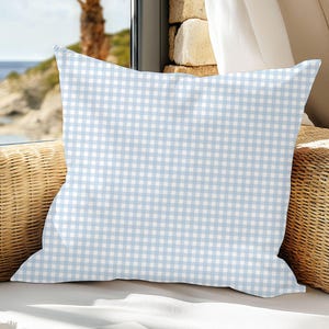 May include: A square pillow with a light blue and white gingham pattern. The pillow is placed on a wicker chair near a window overlooking the ocean. The pillow is a decorative item.
