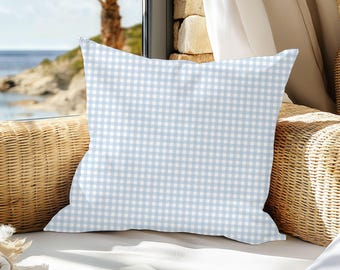May include: A square pillow with a light blue and white gingham pattern. The pillow is placed on a wicker chair near a window overlooking the ocean. The pillow is a decorative item.