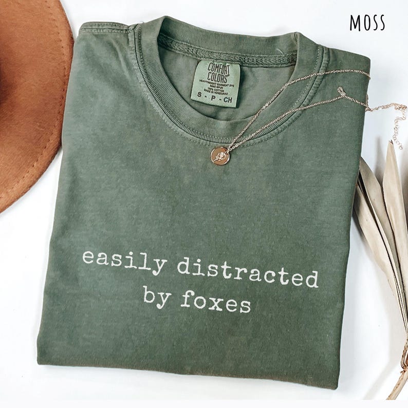 Pu&ograve; includere: T-shirt verde muschio con la scritta bianca "easily distracted by foxes". Una collana d'oro con un piccolo ciondolo &egrave; appoggiata sulla maglietta piegata. La maglietta ha un'etichetta Comfort Colors.