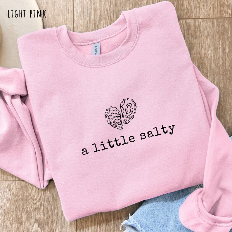 Pode incluir: Camisola rosa claro com a frase "a little salty" em preto, abaixo de um desenho a tra&ccedil;o preto de duas conchas de ostra. As palavras "light pink" est&atilde;o no canto superior esquerdo da imagem.