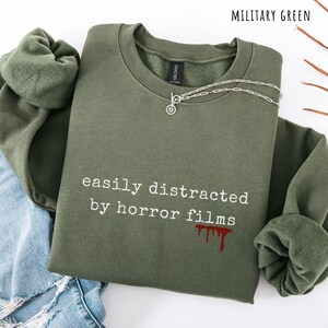 May include: Military green sweatshirt with the text "easily distracted by horror films" in white, and a red blood drip graphic. A silver necklace with a circular pendant is on the sweatshirt. The sweatshirt is folded on top of blue jeans.