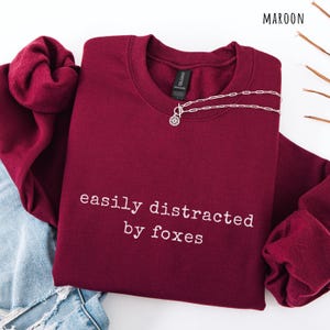 Pu&ograve; includere: Felpa bordeaux con la scritta bianca "easily distracted by foxes". Una collana d'argento con un ciondolo circolare &egrave; sulla felpa. La felpa &egrave; piegata sopra un paio di jeans.