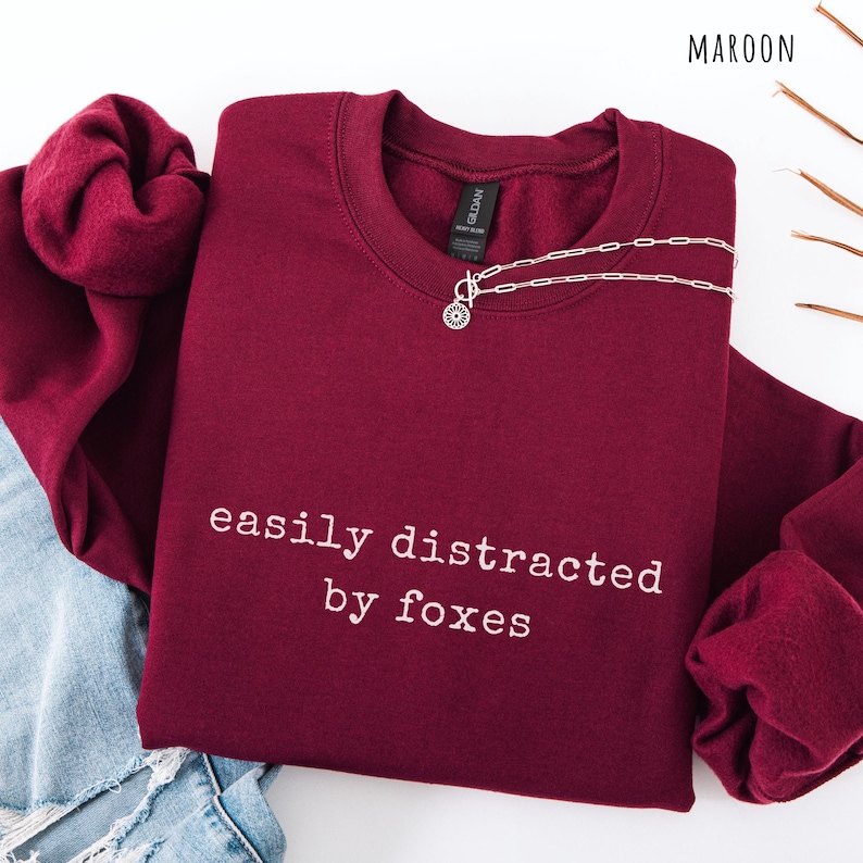 Pu&ograve; includere: Felpa bordeaux con la scritta bianca "easily distracted by foxes". Una collana d'argento con un ciondolo circolare &egrave; sulla felpa. La felpa &egrave; piegata sopra un paio di jeans.