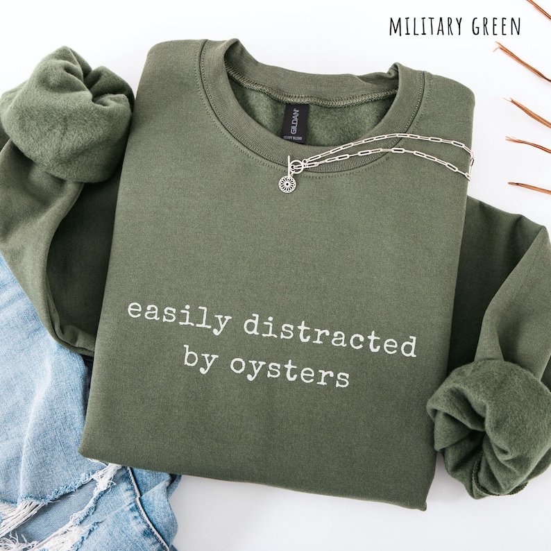 May include: A military green sweatshirt with the text "easily distracted by oysters" in white. A silver necklace with a pendant is on the sweatshirt. The sweatshirt is folded, with a portion of blue jeans visible.