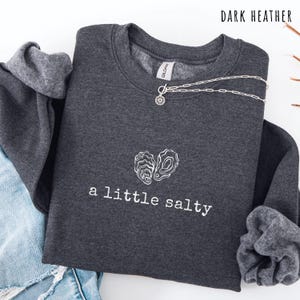 Pode incluir: Camisola cinza escuro com a frase "a little salty" e um desenho de ostra branca. Um colar de prata com um pingente de sol est&aacute; drapeado por cima. As palavras "Dark Heather" est&atilde;o impressas no topo.