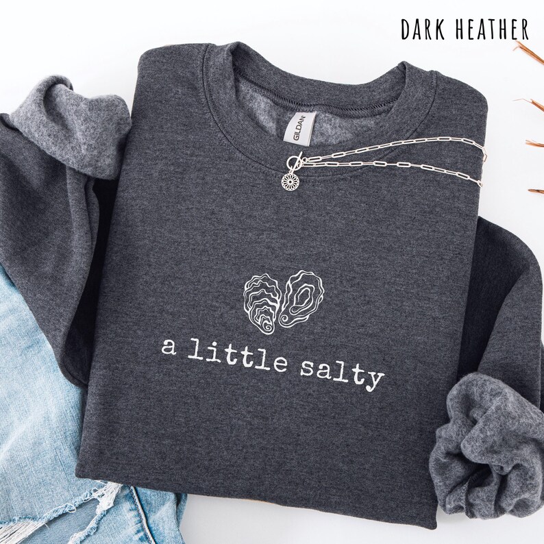 Pode incluir: Camisola cinza escuro com a frase "a little salty" e um desenho de ostra branca. Um colar de prata com um pingente de sol est&aacute; drapeado por cima. As palavras "Dark Heather" est&atilde;o impressas no topo.