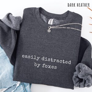 Pu&ograve; includere: Felpa grigio scuro con la scritta bianca "easily distracted by foxes". Una collana d'argento con un ciondolo a forma di sole &egrave; drappeggiata sopra. La felpa &egrave; piegata con del denim visibile.
