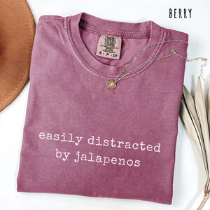 Może przedstawiać: Koszulka w kolorze jagodowym z białym napisem "easily distracted by jalapenos". Koszulka ma okrągły dekolt i kr&oacute;tkie rękawy. Złoty naszyjnik z wisiorkiem jest ułożony na koszulce.