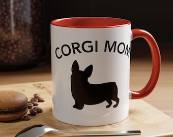May include: White ceramic mug with a red interior and handle. The mug features the text "CORGI MOM" above a black silhouette of a Corgi dog. A spoon and a macaron are on a wooden surface.